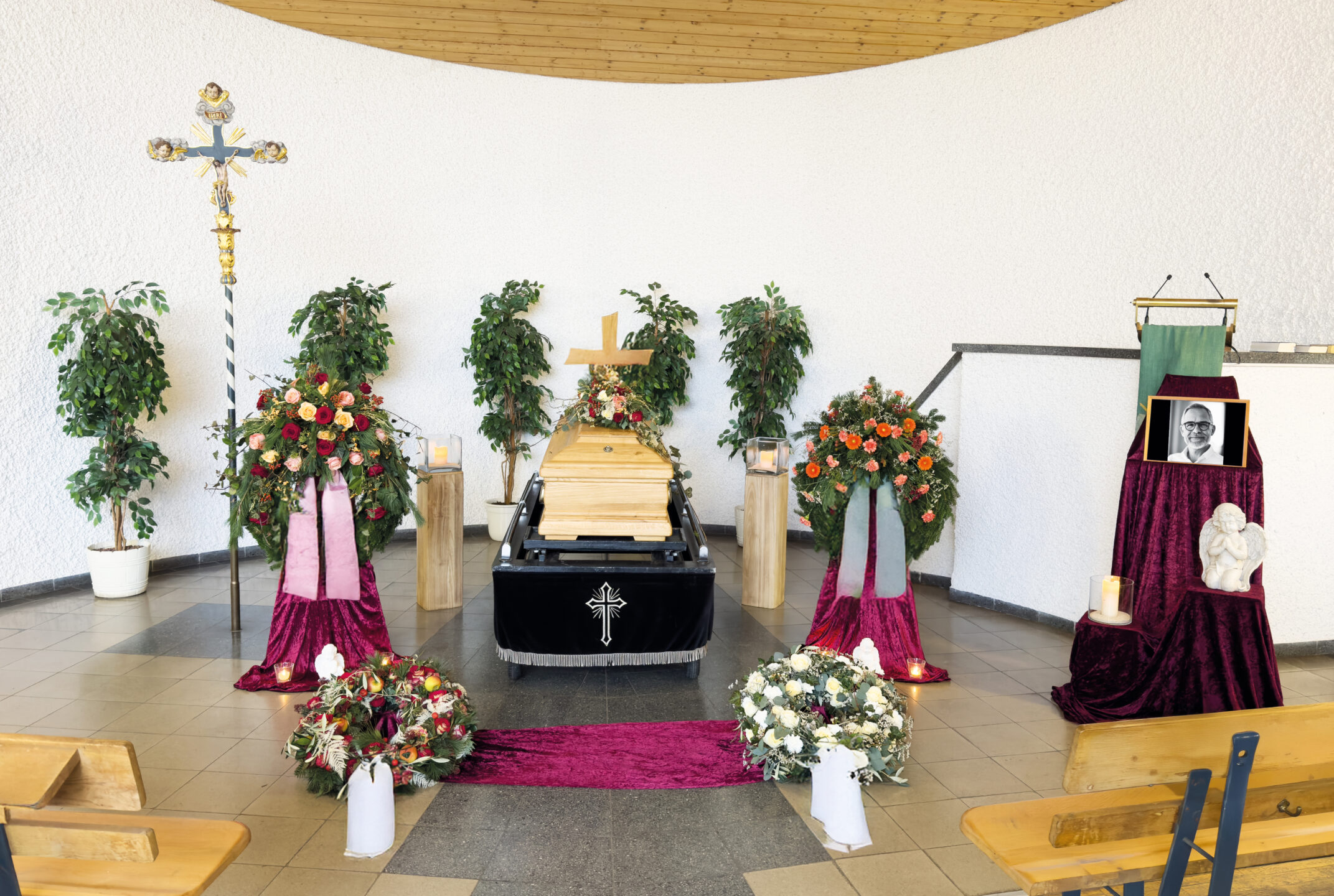 Eine Beerdigungssituation mit einem Holzsarg, auf dem ein Kreuz und Blumen stehen, umgeben von Kränzen, Topfpflanzen und Kerzen. Ein gerahmtes Schwarz-Weiß-Foto und eine weiße Skulptur sind auf einem drapierten Tisch ausgestellt.